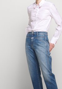 Vit långärmad skjorta med knappränder och röda detaljer, i kombination med ljusblå denimjeans med urblekta sektioner och kontrasterande sömmar.