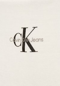 Bílá látka s textem "Calvin Klein Jeans" a výrazným černým logem "CK". Materiál vypadá hladce s jemnou texturou.