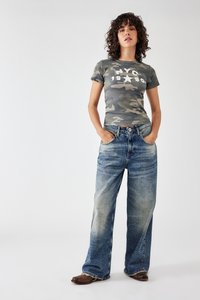Camouflaged t-shirt in grijs en groen met de opdruk "NYC 1980", gecombineerd met blauwe wijde jeans met vervaagde delen en zakken. Bruine schoenen.