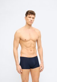 Άνδρας χωρίς πουκάμισο που φοράει σκούρα μπλε αντρικά εσώρουχα με πορτοκαλί μέση με ετικέτα "LACOSTE", στέκεται μπροστά σε απλό φωτεινό φόντο.