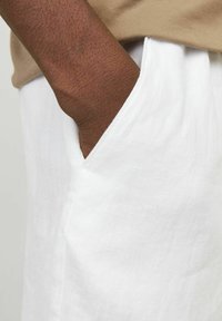 Pantalons en lin blancs avec une poche latérale, tissu texturé et coupe ample, associés à une chemise beige, mettant en avant une tenue décontractée.