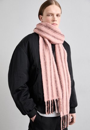 Osoba ubrana w czarną kurtkę i długi, puszysty różowy szalik w paski owinięty wokół szyi, z rękami w kieszeniach, na neutralnym tle.
