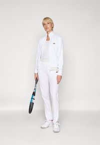 Valge sportlik spordikomplekt, millel on tõmblukuga jope ja püksid, millel on peened triibud. Mudel hoiab tennisereketit ja kannab valgeid sportjalatseid.