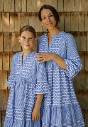 Frau und Mädchen stehen nebeneinander, tragen passende blaue und weiße gemusterte Kleider vor einem Hintergrund aus Holzschindeln.