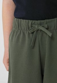 Pantalons vert olive à taille élastique en tissu texturé, avec un détail de nœud à l'avant et une coupe décontractée. La main repose sur le côté.