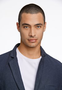 Marinblå herringbone-blazer över en vit t-shirt, med en strukturerad krage och rena linjer, kortklippt hår och neutral bakgrund.