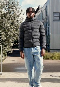Sort pufferjakke med vandret syninger, parret med lyseblå, løstsiddende jeans. Udendørs miljø med grønne omgivelser i baggrunden.