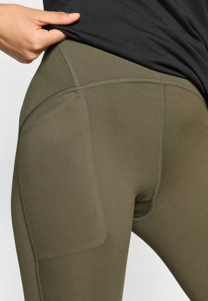 Leggings de color verde oliva hechos de un tejido liso y elástico. Cuentan con un bolsillo lateral y costuras en contraste para un detalle adicional.