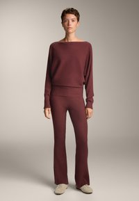 Ribban pletena udobna oblačila v temno burgundski barvi, ki vsebujejo širok pulover z čolnasto izrezom in razširjene hlače z subtilno teksturo ter sproščenim krojem.