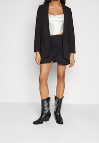 Blazer nero, top corsetto in satin bianco, gonna avvolgente nera con dettagli sui bottoni e stivali cowboy neri al ginocchio con ricami bianchi.