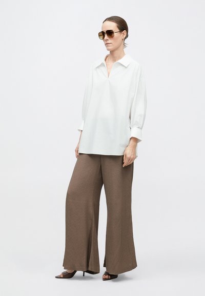 Nő áll, túlméretezett fehér blúzt, bőszárú barna nadrágot, barna, nyitott orrú magassarkú cipőt és nagy napszemüveget visel egyszínű háttér előtt.