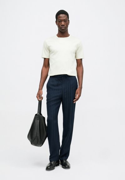 Šviesiai pilkas trumpomis rankovėmis marškinėliai, tamsiai mėlyni dryžuoti kelnės, juodi odiniai batai, juoda rankinė rankoje. Minimalistinis dizainas su subtiliomis tekstūromis.