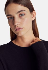 Czarny sweter dzianinowy z okrągłym dekoltem i długimi rękawami. G ładka tekstura z prążkowanymi akcentami na mankietach. Modelka o jasnej karnacji i naturalnym makijażu.