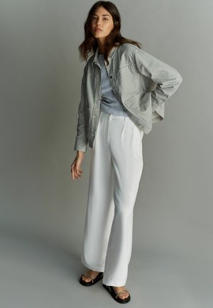 Csíkos, gombos zakó szürke és fehér színben, világoskék felsőre húzva, fehér, bő szárú nadrággal és fekete szandállal, minimális díszítéssel.