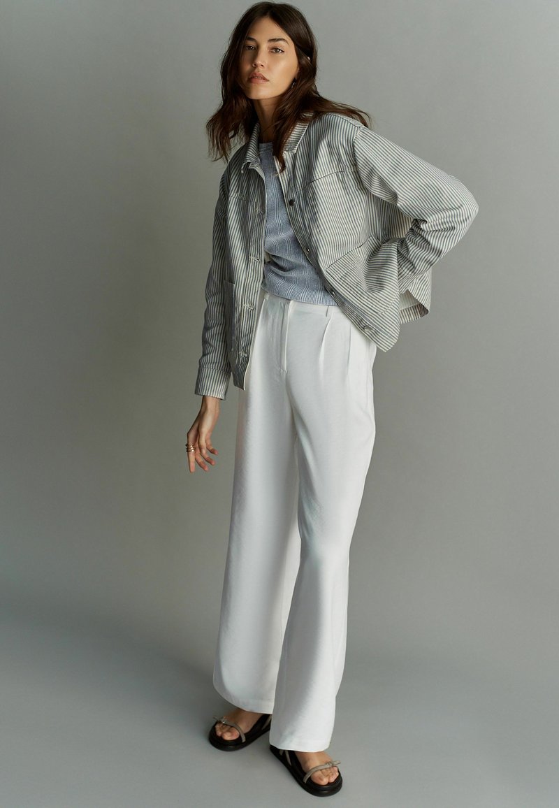Gestreepte, knoopjack in grijs en wit over een lichtblauw top, gecombineerd met witte wijde broek en zwarte sandalen, met een minimale afwerking.