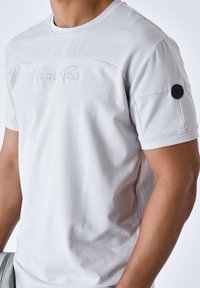 Camiseta gris de manga corta hecha de algodón suave, con detalles de costura, logo bordado en el pecho y un acento circular negro en la manga.