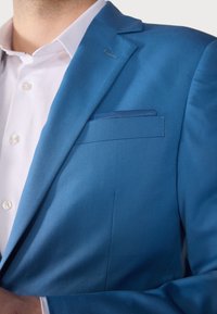 Sinine bleiser, millel on sile tekstuur, ühekordne rinnatasku ja teravad kraed. Kantud valge särgi üle, millel on nööpdetailid.