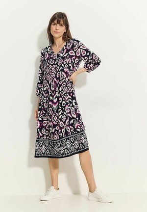 Schwarz-rosa gemustertes Kleid mit V-Ausschnitt, 3/4-Ärmeln und dekorativem Saum. Getragen mit weißen Turnschuhen, locker sitzend und mit weichem Material.