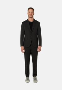 Costume noir à coupe ajustée, veste à simple boutonnage et pantalon assorti. Porté avec une chemise noire et des baskets grises.