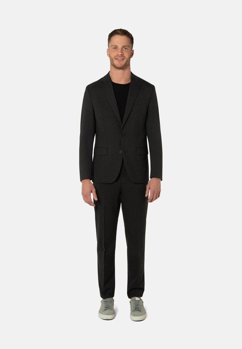 Costume noir à coupe ajustée, veste à simple boutonnage et pantalon assorti. Porté avec une chemise noire et des baskets grises.