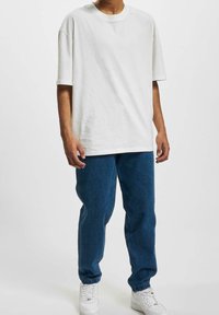 Camiseta de algodón blanca con un corte holgado y mangas cortas, combinada con pantalones de mezclilla azules. Ambas prendas tienen un estilo relajado, con zapatillas blancas.