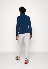 Veste en polaire marine à col haut et coutures bleues contrastées, associée à un pantalon cargo beige clair et des chaussures de sport gris-orange.