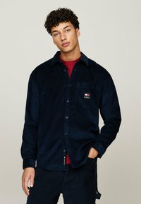 Chemise en velours côtelé bleu marine avec une poche à l'avant, un design à boutons et un petit patch logo. Manches longues avec un col classique et un tissu texturé.