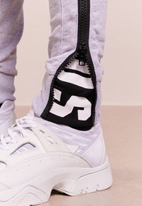 Grå jogger med siddragkedjor som avslöjar svart tyg under, med djärva vita grafiker. Matchad med vita sportskor. Slät textur.