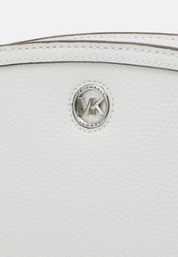 Bílá kožená kabelka s texturovaným povrchem a stříbrným kruhovým logem s vyrytými písmeny "MK" a "MICHAEL KORS".