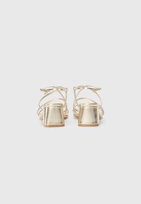 Goldene, metallische Sandalen mit Riemen und Blockabsatz, die eine offene Rückseite und ein Kreuzdesign im oberen Bereich für zusätzliche Details aufweisen.