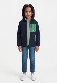 Νεαρό παιδί με κοτσίδες που φοράει μαύρο και πράσινο τζάκετ με φερμουάρ, γκρι μπλουζάκι, μπλε τζιν, γκρι κάλτσες και λευκά αθλητικά παπούτσια, όρθιο με το πρόσωπο προς τα εμπρός.