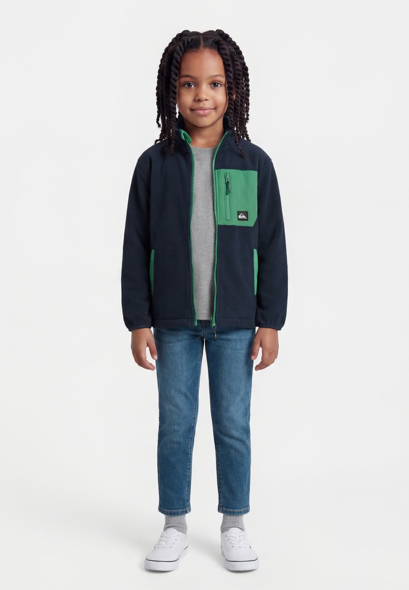 Νεαρό παιδί με κοτσίδες που φοράει μαύρο και πράσινο τζάκετ με φερμουάρ, γκρι μπλουζάκι, μπλε τζιν, γκρι κάλτσες και λευκά αθλητικά παπούτσια, όρθιο με το πρόσωπο προς τα εμπρός.