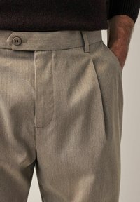 Lichtgrijze geplooide broek van gestructureerde stof. Beschikt over een platte voorkant, knoopsluiting en zijzakken.