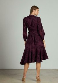 Rochie închisă la culoare, de un violet profund, cu mâneci lungi tip puff, dintr-un material texturat și cu un tiv evazat. Prezintă o talie legată și un model delicat.