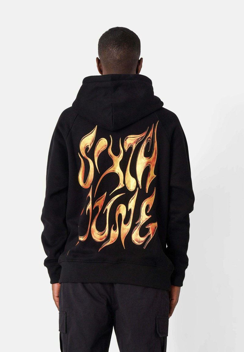 Sudadera negra con un gran diseño gráfico en un texto dorado estilo llama en la parte de atrás. Tiene un bolsillo canguro y puños de canalé.