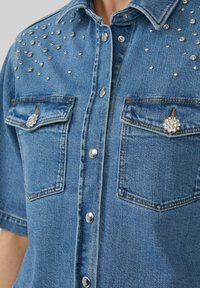 Blauwe denim overhemd met zilveren studs op de schouders, twee borstzakken met decoratieve knopen, korte mouwen, en zilveren drukknoopjes aan de voorkant.