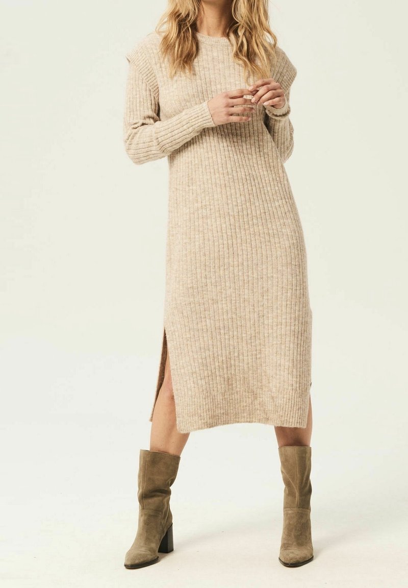 Robe en maille côtelée beige avec manches longues, fentes latérales et coupe décontractée. Associée à des bottes à talons vert olive. Texture douce.