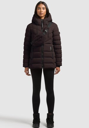 Une femme porte une doudoune à capuche marron foncé, un sac en polaire en bandoulière, un legging noir et des bottes d’hiver sur un fond gris uni.