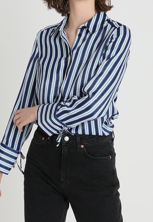 Person wearing a long-sleeve, vertical blue, black, and white striped shirt tucked into high-waisted black jeans with button and pockets.