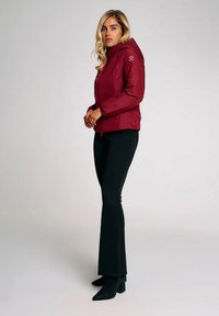 Donna bionda in piedi con pantaloni neri, stivali con tacco neri e una giacca con cappuccio bordeaux, su uno sfondo chiaro e uniforme.