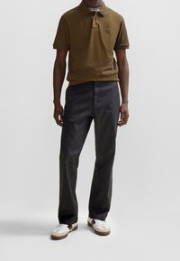 Polo vert olive avec col et logo sur la poitrine, associé à un pantalon noir droit et des baskets blanches avec des accents noirs.