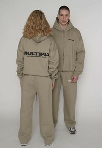 Zwei Models tragen beige Kapuzenpullover und Hosen mit dem Schriftzug "MULTIPLY", eines zeigt die Vorderseite, das andere die Rückseite.
