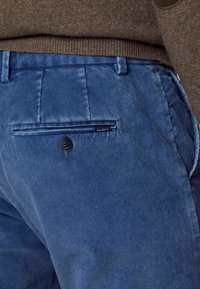 Pantalon en velours côtelé bleu avec une poche arrière boutonnée et ourlet de pull brun visible au-dessus de la taille.