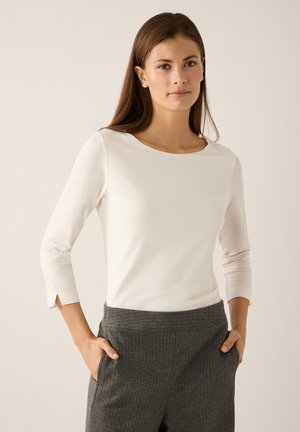 Wit shirt met lange mouwen, een ronde halslijn en schuine manchetten, gecombineerd met grijze pantalon met krijtstrepen en zijzakken.