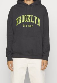 Sudadera con capucha de color gris carbón hecha de tela, con un gran gráfico en neón amarillo que dice "BROOKLYN USA 2007", con un bolsillo delantero y capucha con cordón.