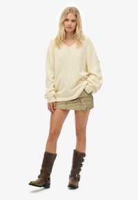 Blondynka stoi z rękami w kieszeniach, nosząc oversize'owy kremowy sweter, beżową mini spódnicę z kieszeniami oraz brązowe botki na klamrę.