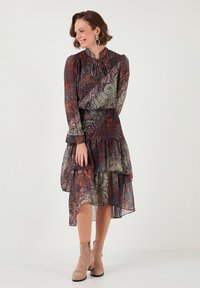 Langärmliges, gemustertes Kleid in dunklen Farbtönen mit gerafftem Saum. Durchscheinender Stoff, tailliert und mit hohem Kragen. Kombiniert mit beigen Ankle Boots.