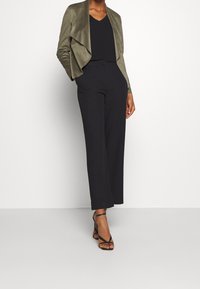 Giacca in suede verde oliva con frontale drappeggiato, top nero a scollo a V, pantaloni neri a gamba larga e tacchi neri con cinturini. Posata su uno sfondo bianco.