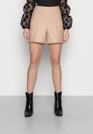 Frau trägt beige maßgeschneiderte Shorts, ein schwarzes Top mit transparenten gepunkteten Puffärmeln und schwarze Knöchelstiefel, steht auf grauem Boden.