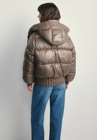 Puchowy brązowy kurtka puchowa z kapturem, z pikowanym wzorem, ściągaczami przy rękawach i elastycznym dołem, zestawiona z niebieskimi dżinsami.